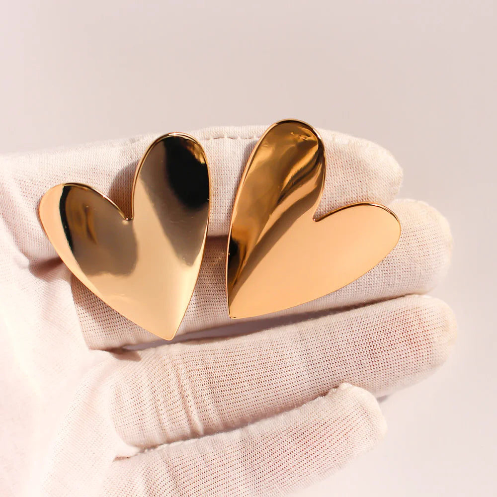 Large polished gold heart stud earrings held on a white glove